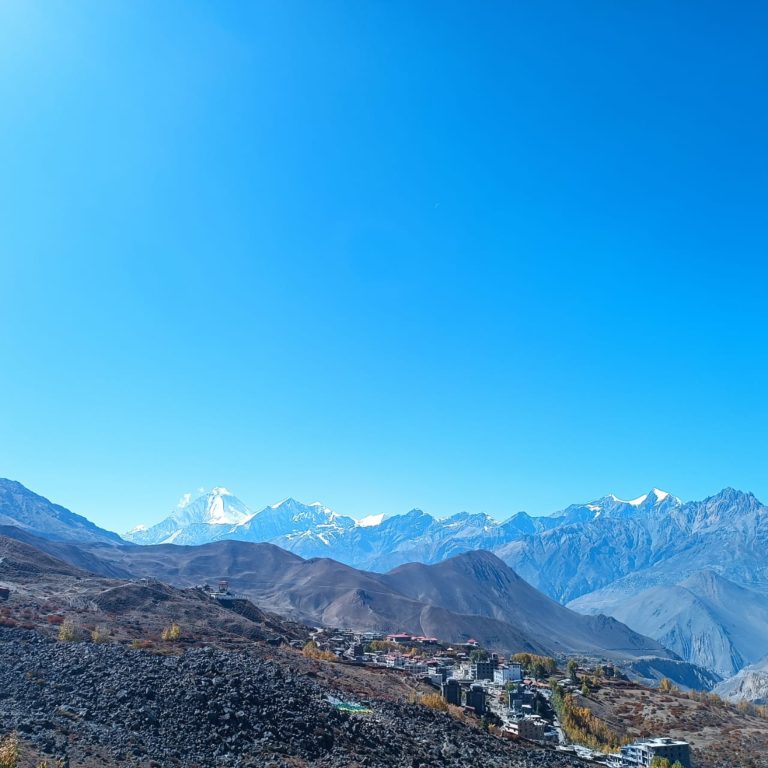 A panoramic view of a rugged mountain landscape with snow-capped peaks under a clear blue sky. In the foreground, there is a small, scattered settlement nestled among rolling hills and rocky terrain.