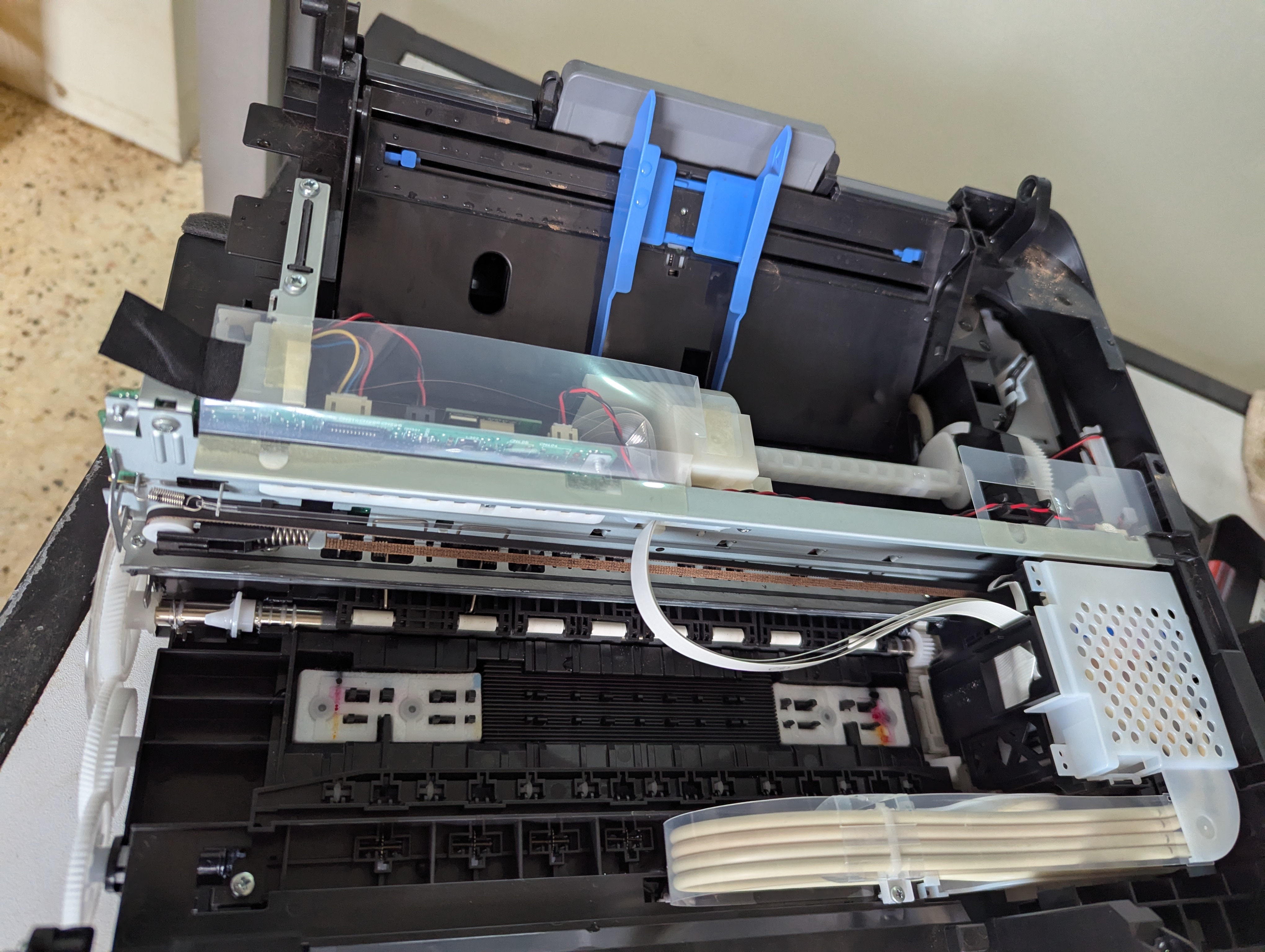 The image shows the internal components of an open printer or copier. Various mechanical and electronic parts are visible, including gears, wires, and connectors. The machine appears to be disassembled, revealing its intricate inner workings.