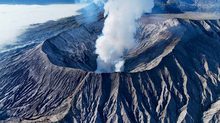 Aerial view of Mount Bromo’s crater with thick smoke rising from its depths, showcasing the rugged volcanic terrain and steep crater walls.