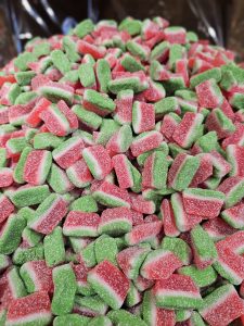 A pile of watermelon-shaped gummy candies. The candies are shaped like small, thick slices of watermelon with a green rind, white layer, and red center with tiny speckles.