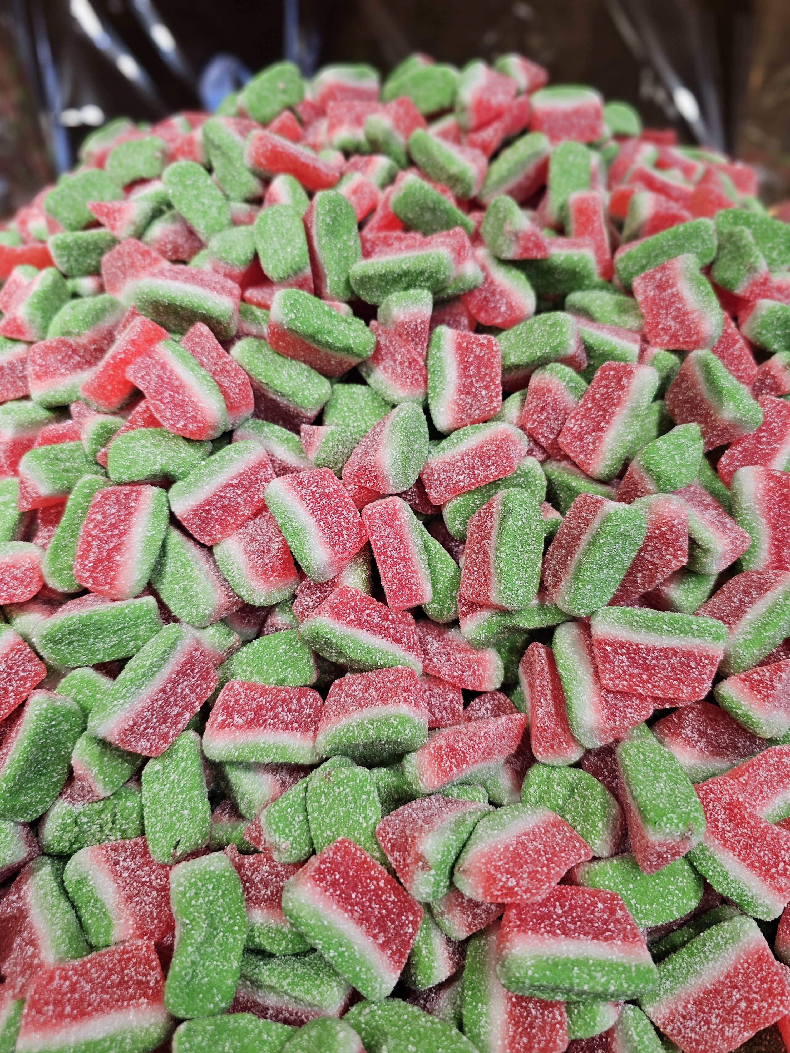 A pile of watermelon-shaped gummy candies. The candies are shaped like small, thick slices of watermelon with a green rind, white layer, and red center with tiny speckles.