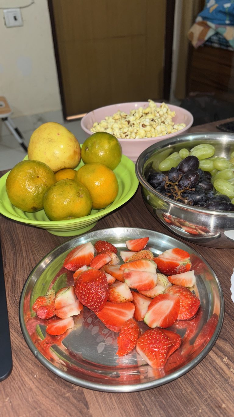 A table displaying a bowl of strawberries, oranges, and grapes alongside a bowl of popcorn.