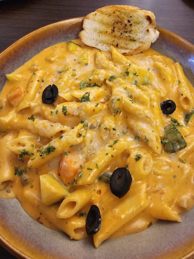 A plate of penne pasta coated in a creamy orange sauce, garnished with black olives and parsley and accompanied by a slice of garlic bread. The dish is presented on a speckled beige plate with a slightly raised edge, set against a dark wooden table.