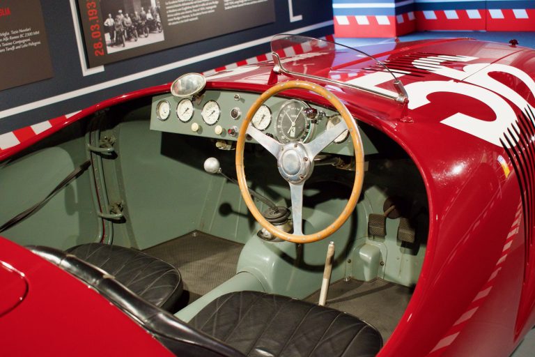 Interior of a vintage red racing car, featuring a wooden steering wheel, multiple dashboard gauges, two black leather seats, and a minimalist gear shift.