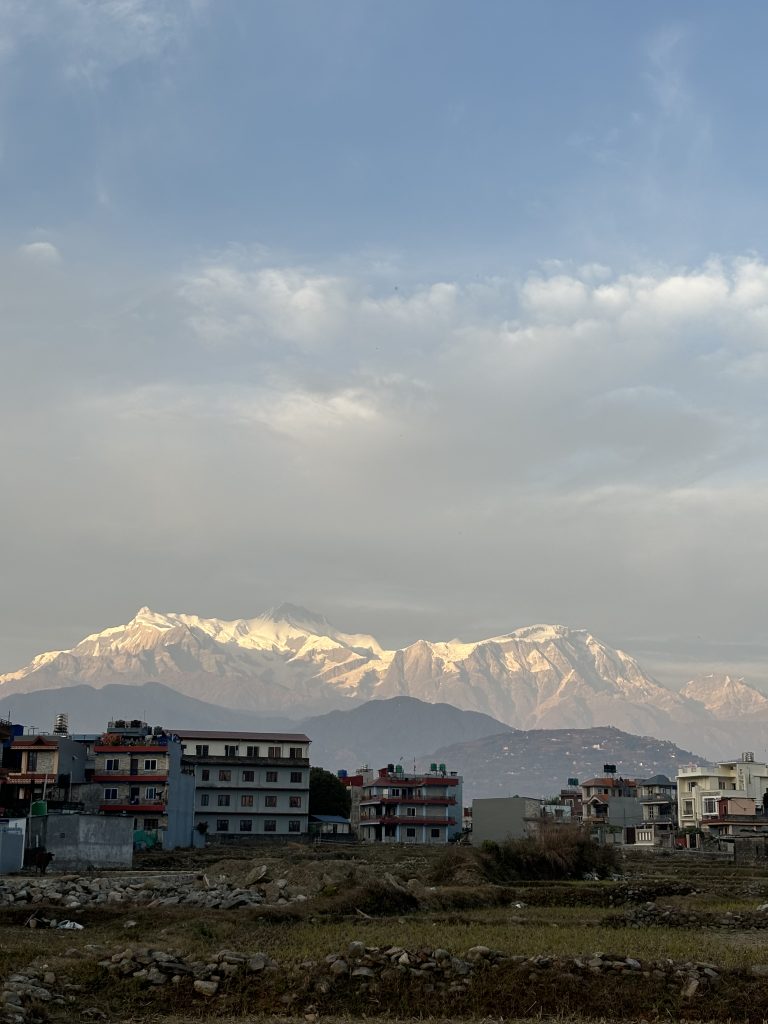 A scenic view of snow-capped mountains under a partly cloudy sky, with a foreground of residential buildings and open fields.