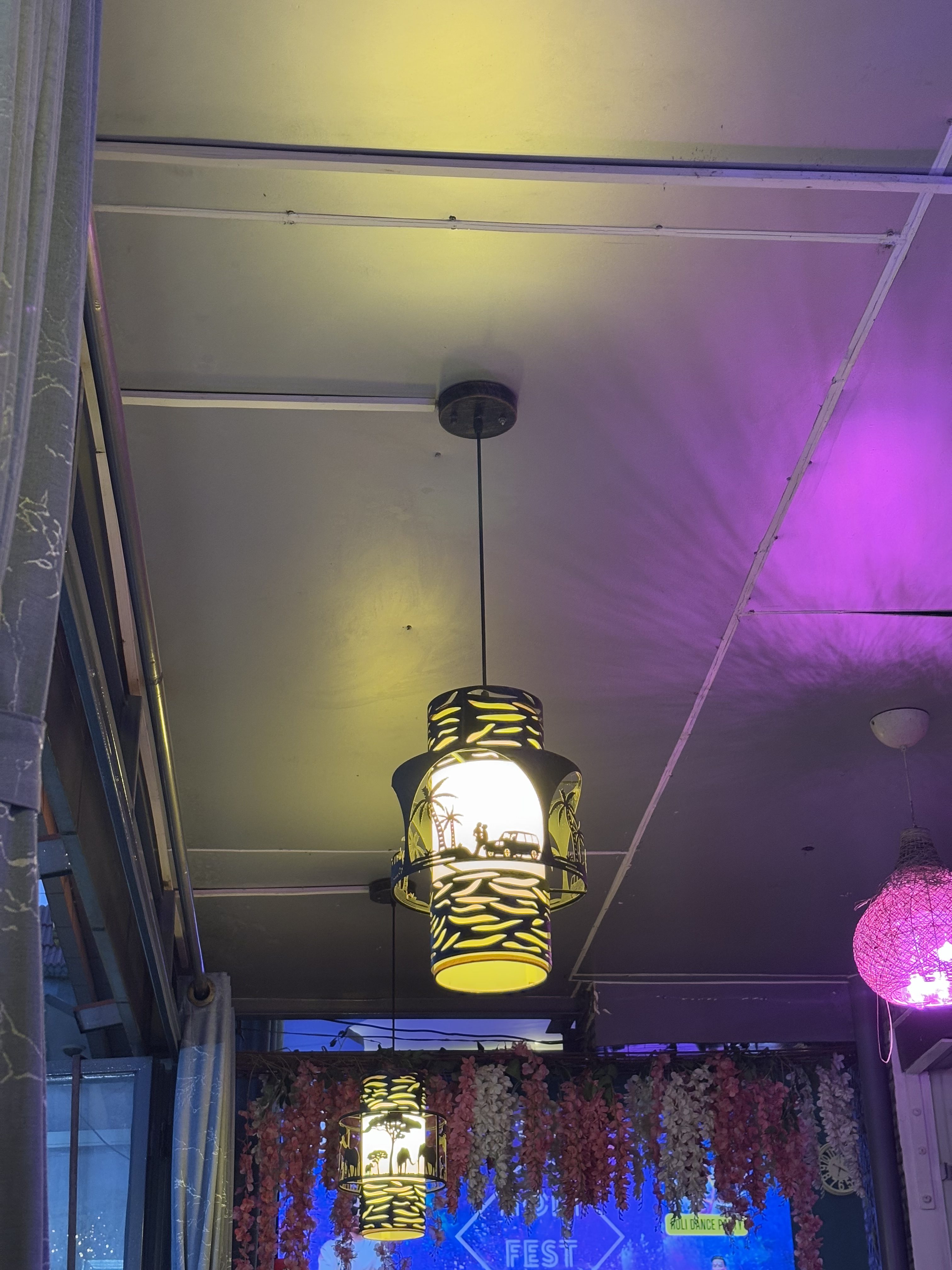 Hanging pendant lights with intricate cutout designs featuring animal silhouettes and trees, illuminating a room with colorful lighting. Decorative flowers hang in the background, and there is a wall with a sign partially visible at the bottom.