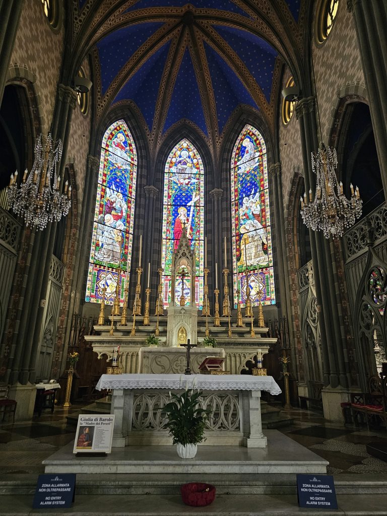 The interior of the Chiesa di Santa Giulia in Torino, Italy, features the altar with a white cloth and a potted plant in front, three stained glass windows behind the altar, and two chandeliers hanging from the ceiling.