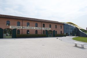 Exterior view of the Enzo Ferrari Museum in Modena, featuring a historic red-brick building labeled “Officina Meccanica Alfredo Ferrari” and a modern, curved glass extension with a yellow roof.
