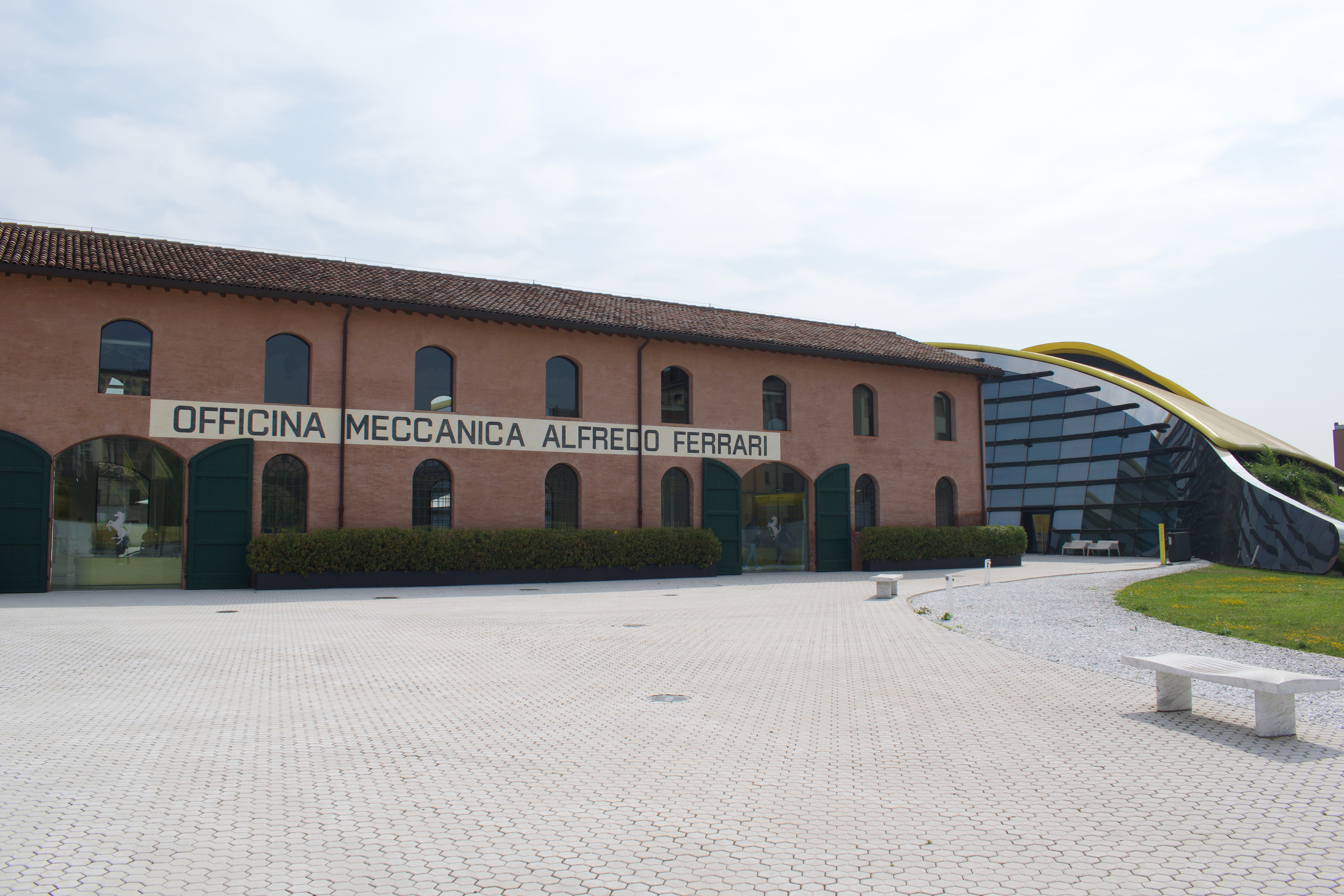 Exterior view of the Enzo Ferrari Museum in Modena, featuring a historic red-brick building labeled "Officina Meccanica Alfredo Ferrari" and a modern, curved glass extension with a yellow roof.