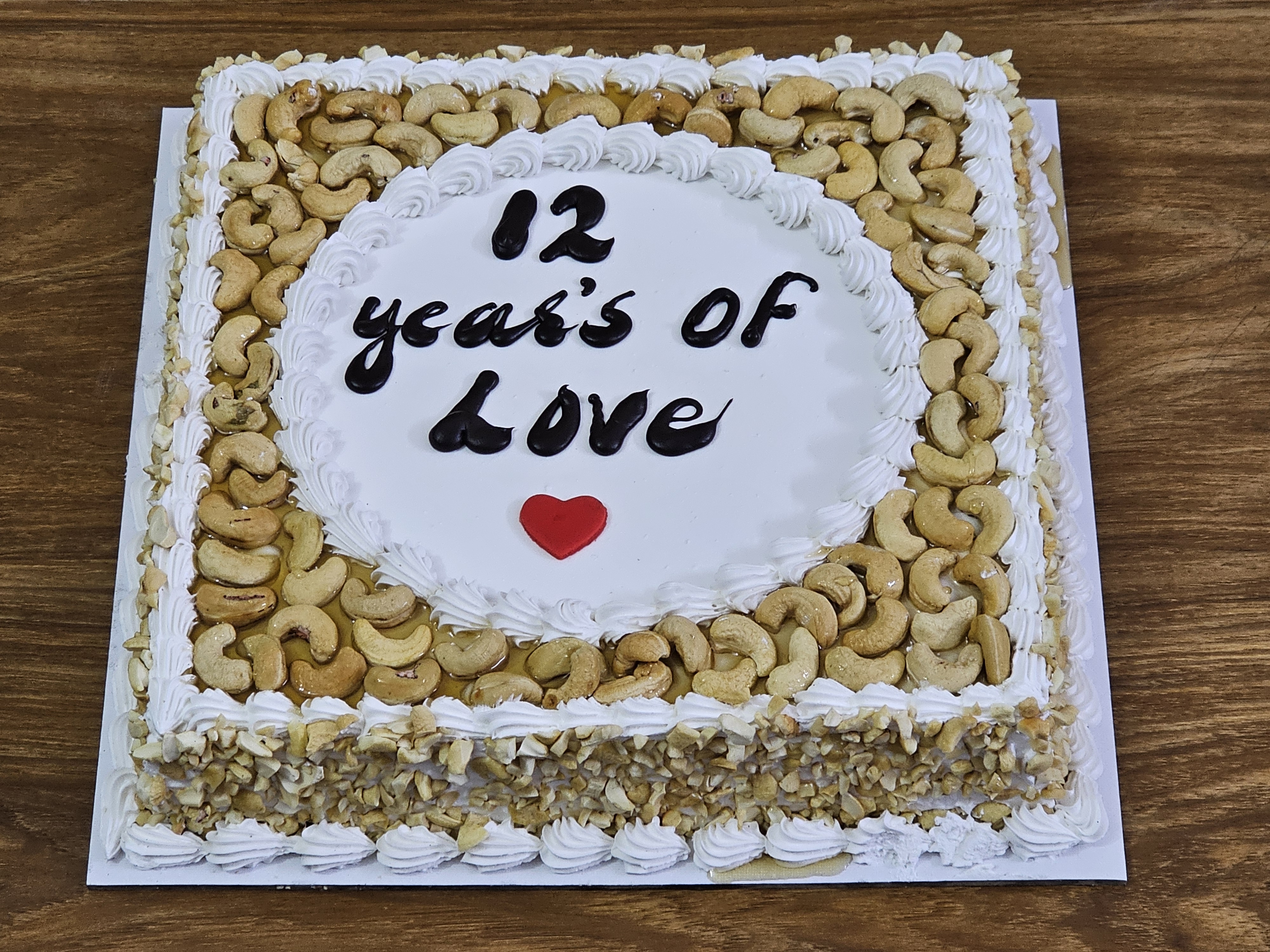 A square cake decorated with cashews and frosting, with the inscription "12 years of Love" written in black icing and a small red heart underneath. The cake is placed on a white square base and set against a wooden surface. Captured from Perumanna, Kozhikode.