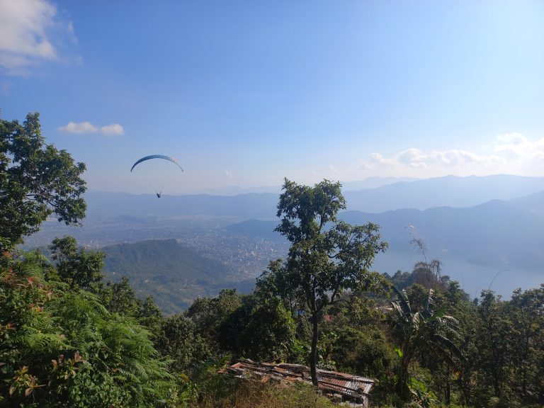 A paraglider soars in the sky over a lush, mountainous landscape with a city and a lake visible below. The foreground includes dense green foliage and a small structure with a corrugated metal roof. The sky is clear with a few clouds and the scene is set against distant mountain ranges.