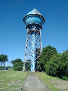 A large blue water tower with an intricate metal support structure stands against a clear blue sky. The surrounding area features green grassy lawns and trees, with a pathway leading towards the tower. A car is visible on a road to the left.