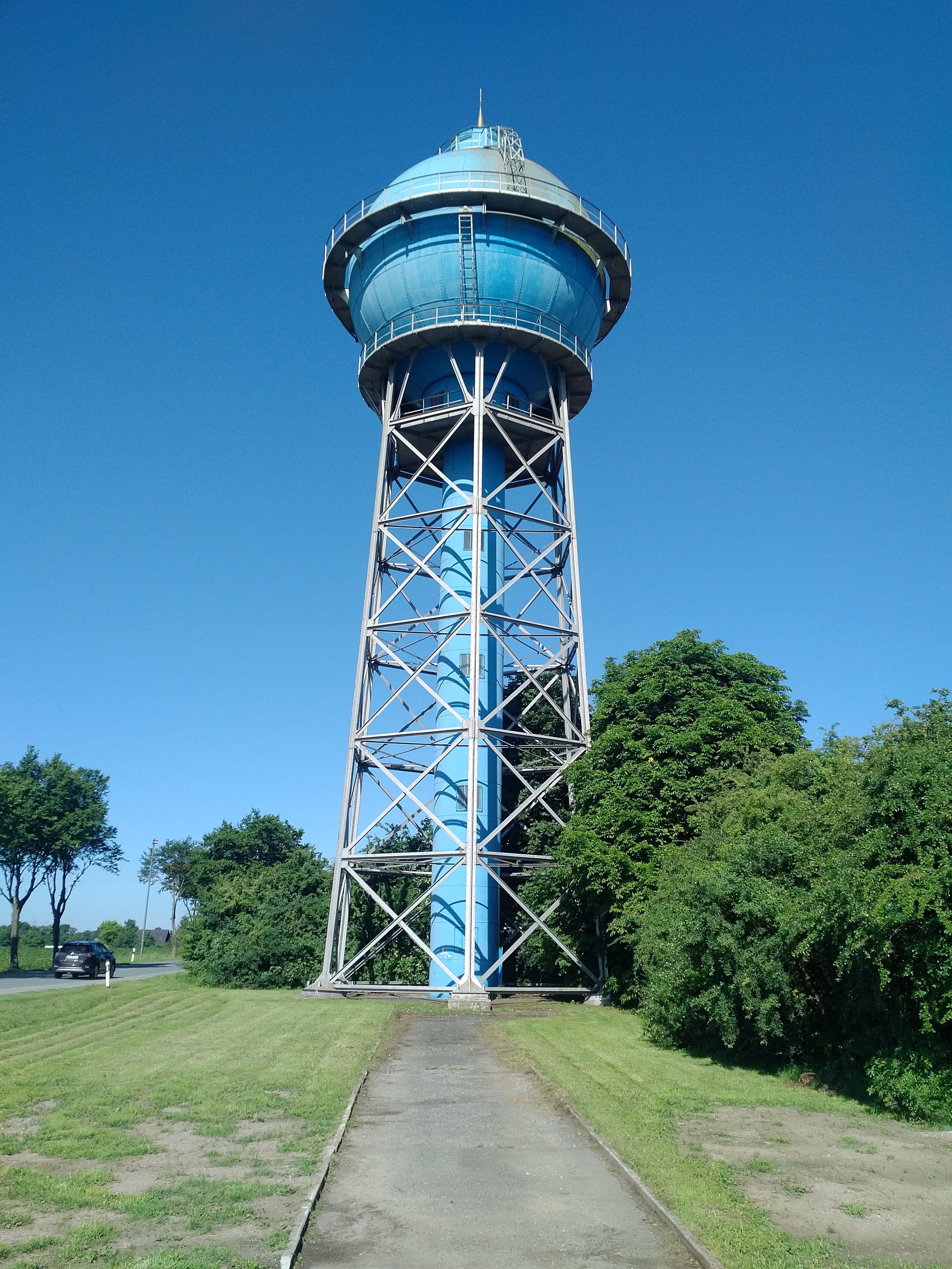 A large blue water tower with an intricate metal support structure stands against a clear blue sky. The surrounding area features green grassy lawns and trees, with a pathway leading towards the tower. A car is visible on a road to the left.