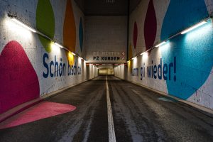 A tunnel with colorful painted walls featuring large colored shapes and German text. The sides are illuminated by fluorescent lights, and there's a road with a single white line running through the center.