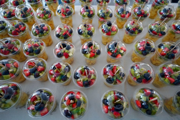 Rows of plastic cups filled with assorted fresh fruit, including strawberries, blueberries, and kiwis, covered with clear lids and arranged neatly on a table.