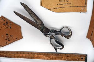 A pair of large, vintage metal scissors is placed on a white surface, surrounded by pattern templates and a wooden ruler. The templates have sewing instructions and measurements printed on them.

