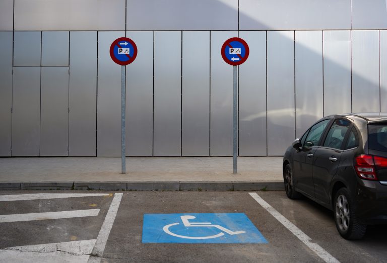 A parking area in front of a metallic wall features a parking spot with a blue disabled sign on the ground. Two signs on poles indicate parking instructions, and a dark gray car is partially visible, parked in the adjacent space.