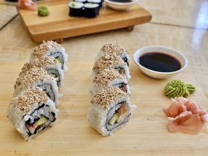 A wooden board with California rolls, soy sauce, wasabi, and pickled ginger. The California rolls are topped with sesame seeds and arranged in two rows. Additional sushi pieces are visible on a separate wooden board in the background. 
