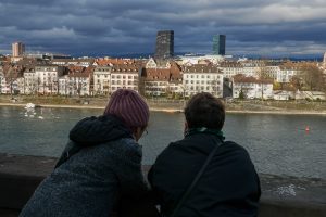 Two people leaning on a stone railing, looking across Rhine river towards Basel, a Swiss city with historic buildings and modern skyscrapers under a cloudy sky.