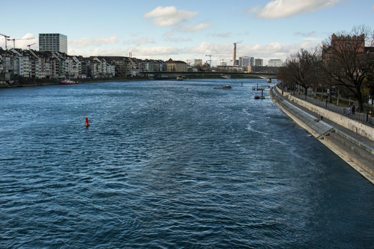 View of the Rhine river passing through Basel, lined with bare trees and a pedestrian walkway. The left side is bordered by a row of buildings, including residential apartments and taller structures.