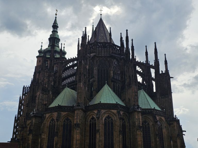 The exterior of St. Vitus Cathedral or Prague Castle, featuring its Gothic architecture with spires, arches, and green rooftops, set against a cloudy sky.