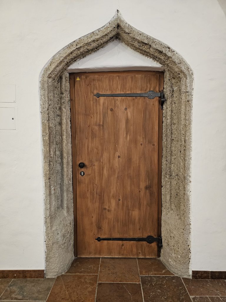 A wooden door set within an arched stone frame. The door is made of vertical wooden planks and has two black metal hinges, one near the top and one near the bottom. A simple black doorknob and keyhole are located on the left side of the door. From Fortress Hohensalzburg, Salzburg.