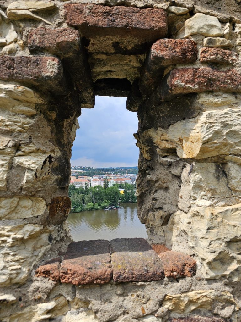 A long view of Prague through a stone-framed opening, part of the Vy?ehrad fortress. The opening is constructed from rough-hewn stones and red bricks, giving it a rustic, aged appearance. Through the opening, the Vltava River can be seen flowing through the city, with buildings and greenery lining its banks.