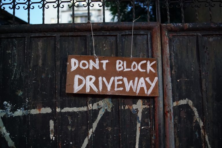 A handwritten sign on a piece of cardboard reading “DON’T BLOCK DRIVEWAY” is attached to a rusty, textured metal gate with wire. In the background, parts of a building and some trees are visible.