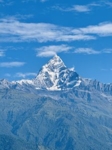 

A majestic snow-capped fishtail (Machapuchare) mountain peak under a clear blue sky with scattered clouds, surrounded by wooded hills.