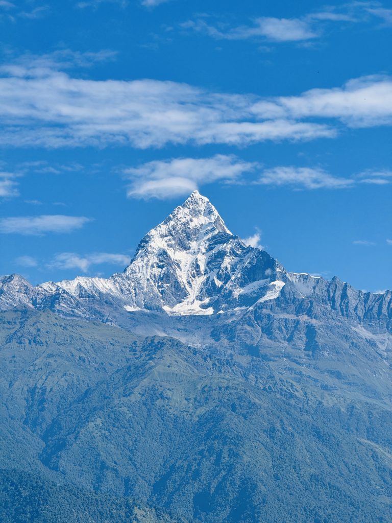 A majestic snow-capped fishtail (Machapuchare) mountain peak under a clear blue sky with scattered clouds, surrounded by wooded hills.