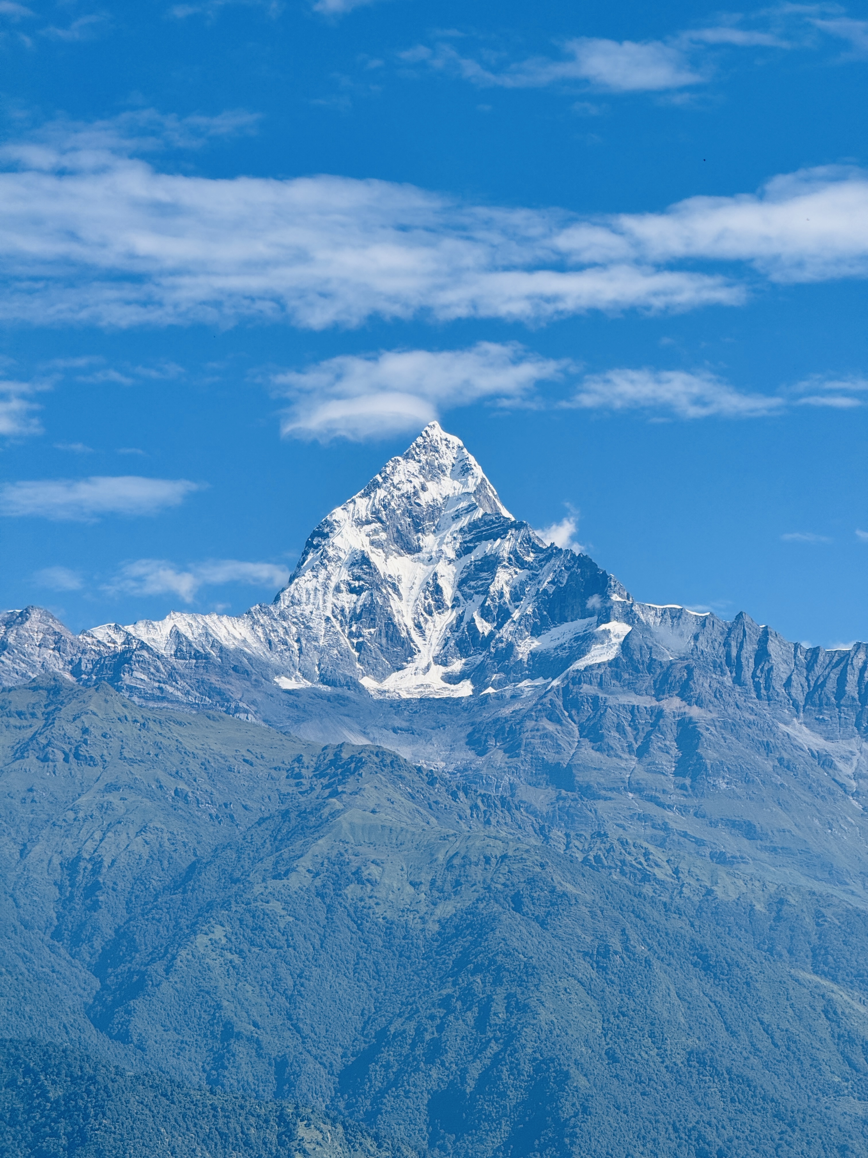 

A majestic snow-capped fishtail (Machapuchare) mountain peak under a clear blue sky with scattered clouds, surrounded by wooded hills.