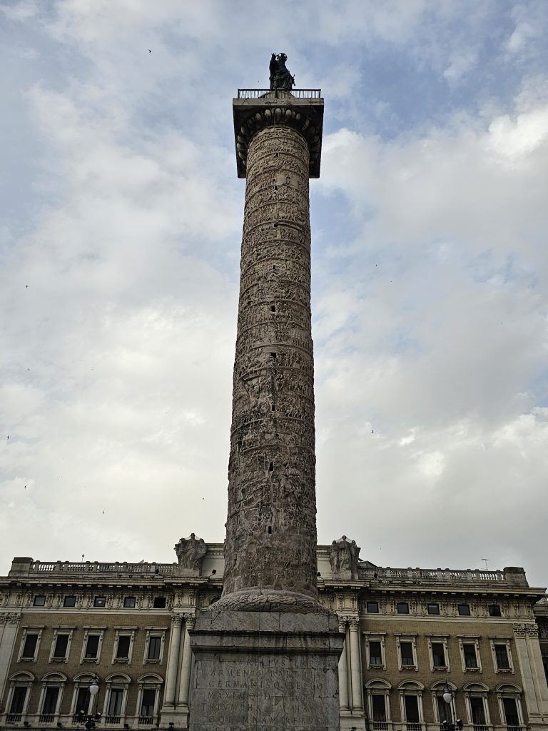 The Column of Marcus Aurelius, a large Roman monument with detailed carvings spiraling around its shaft, is set against a cloudy sky in the evening. The column is positioned in front of a classical building, in the Piazza Colonna in Rome.