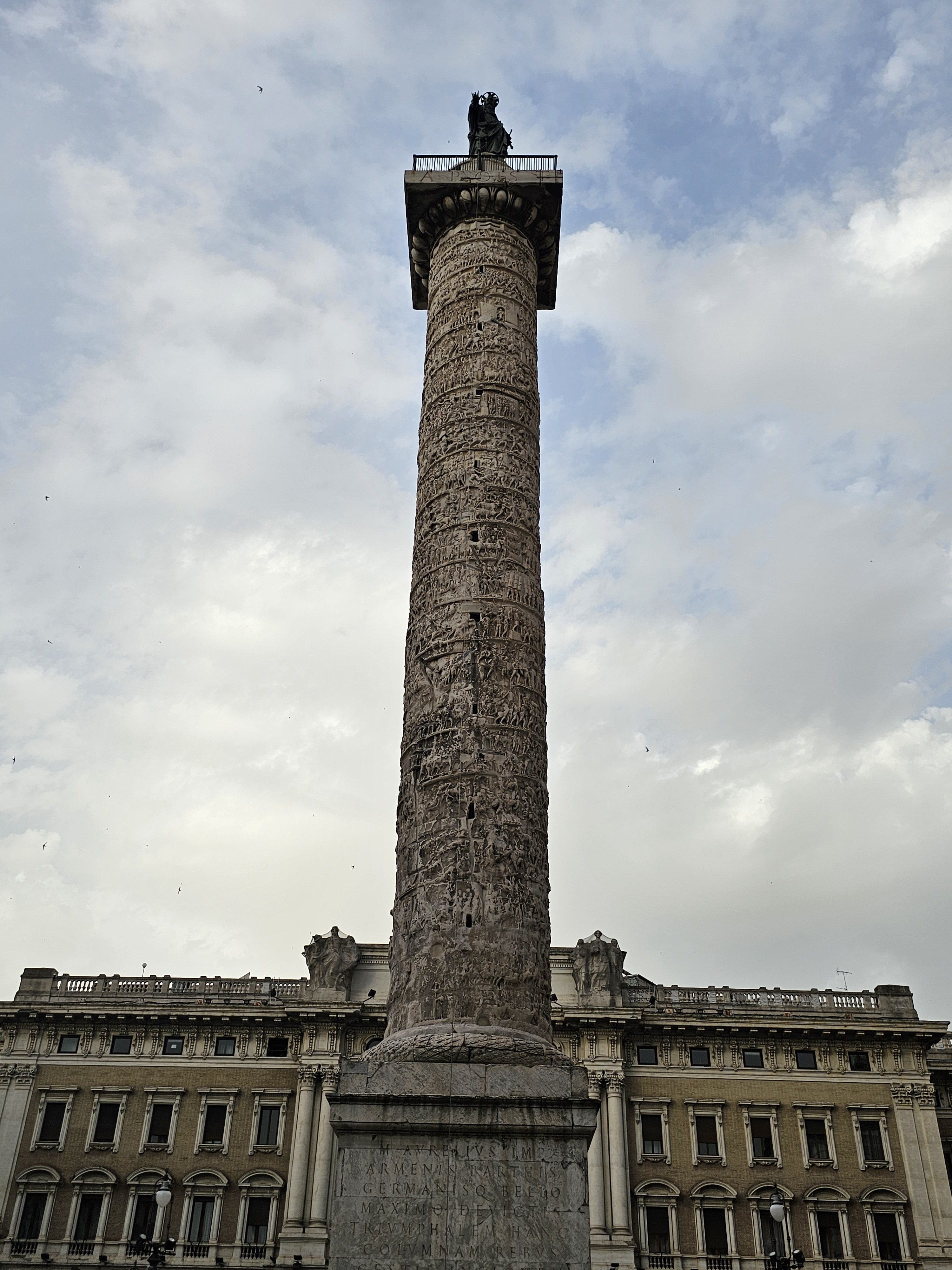 The Column of Marcus Aurelius, a large Roman monument with detailed carvings spiraling around its shaft, is set against a cloudy sky in the evening. The column is positioned in front of a classical building, in the Piazza Colonna in Rome.