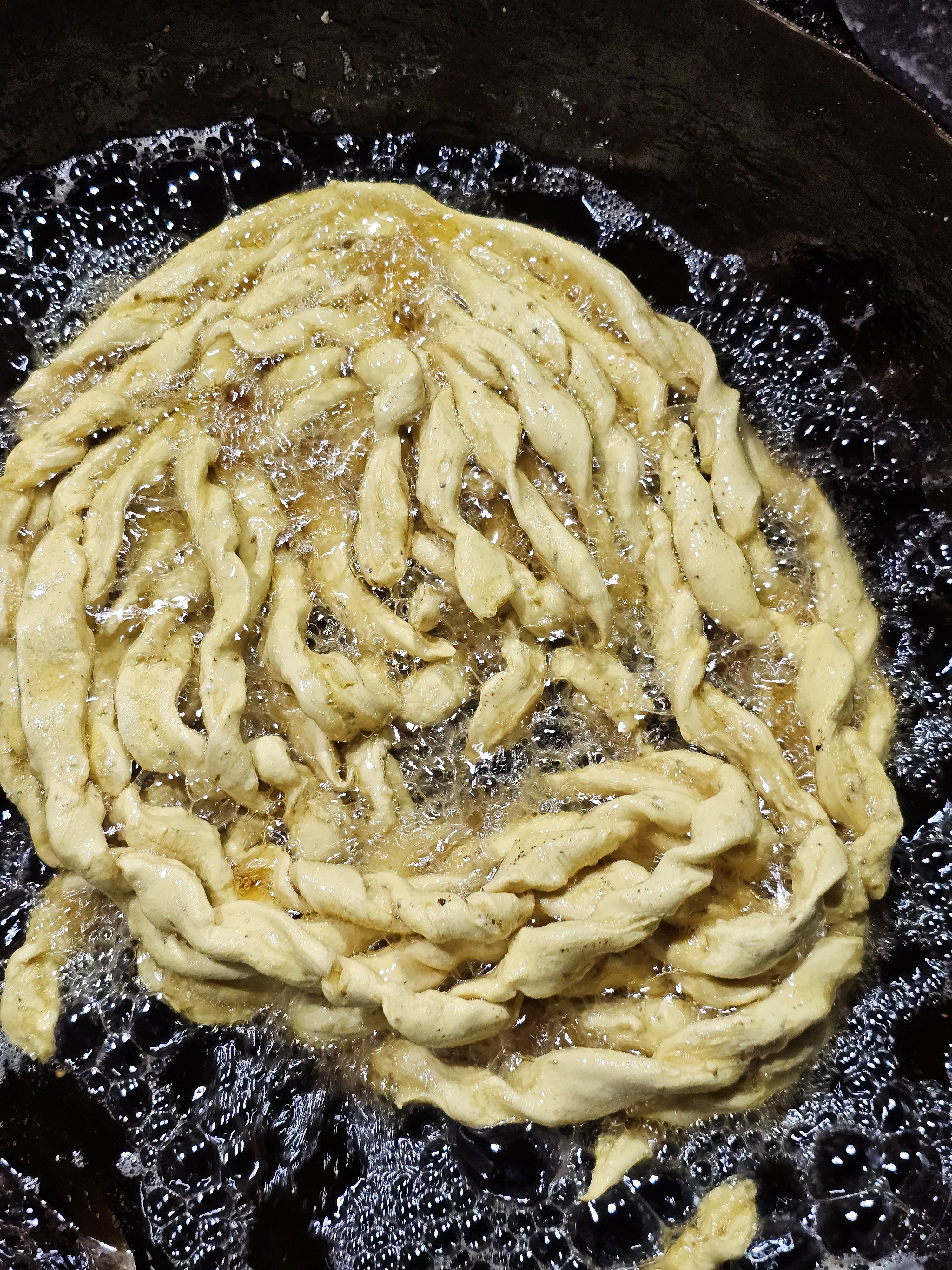 A close-up of Vanela Gathiya is deep-fried in a large, round pan. The snack is golden-brown in color and has a twisted, rope-like shape, it is surrounded by bubbling oil and it is being cooked. Vanela Gathiya is a popular Indian snack, especially in Gujarat, and is known for its soft and fluffy texture. Captured from the streets of Ahmedabad.