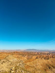 Expansive desert landscape with rugged, eroded hills and valleys under a clear blue sky. Distant mountains can be seen on the horizon.