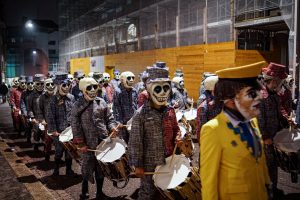 A group of people wearing matching patterned costumes and skull masks, each holding drums and drumsticks, parades through a dimly lit street with construction barriers in the background during Fasnacht in Basel