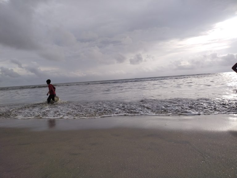 A person walks along the shoreline of a beach, with waves gently crashing and a cloudy sky in the background.