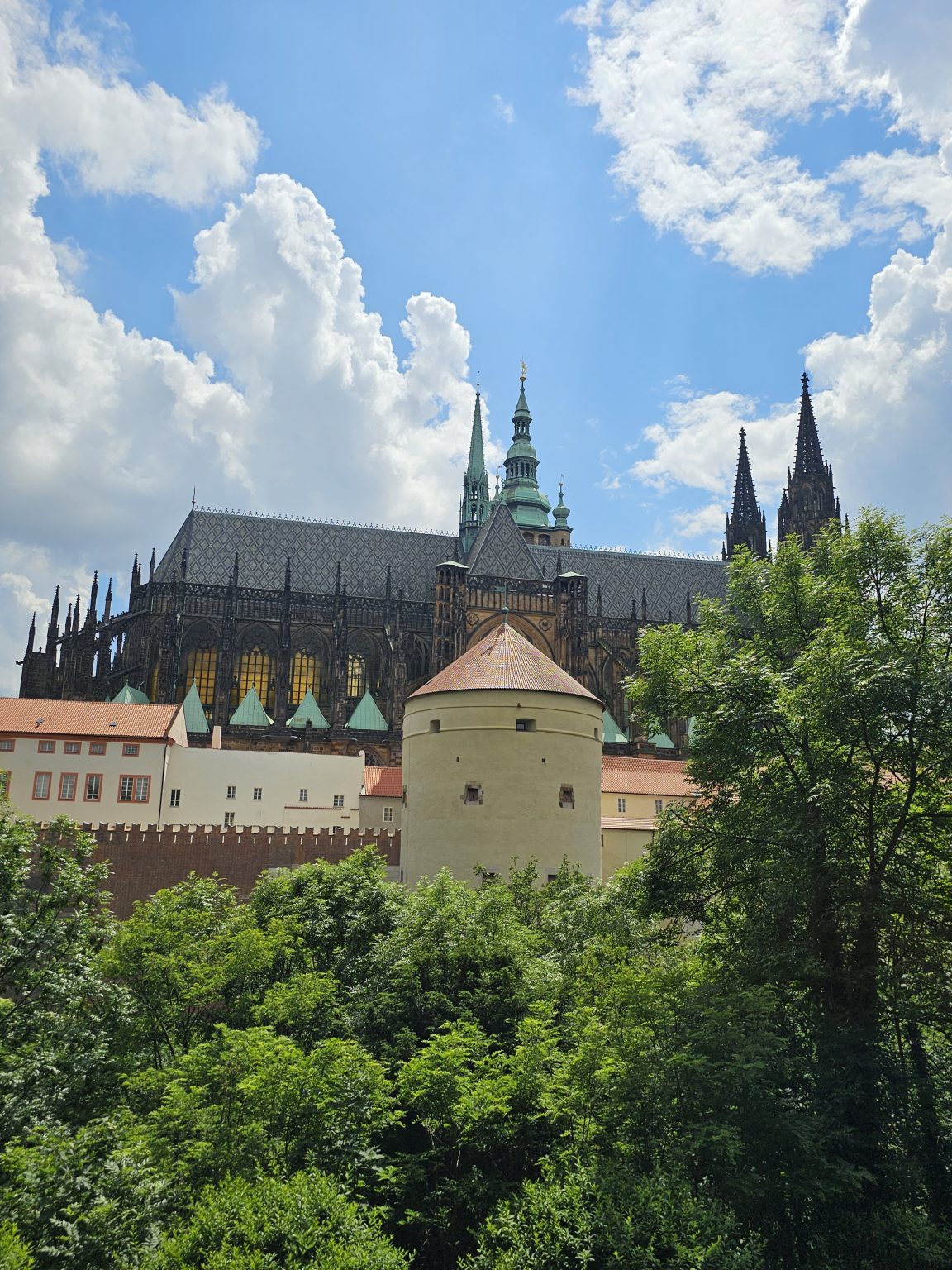 A long view of St. Vitus Cathedral, part of Prague Castle, has a clear blue sky dotted with fluffy white clouds in the background. The cathedral's spires and intricate architectural details are showcasing its Gothic style. In the foreground, a round tower with a red-tiled roof stands out, surrounded by lush green trees.