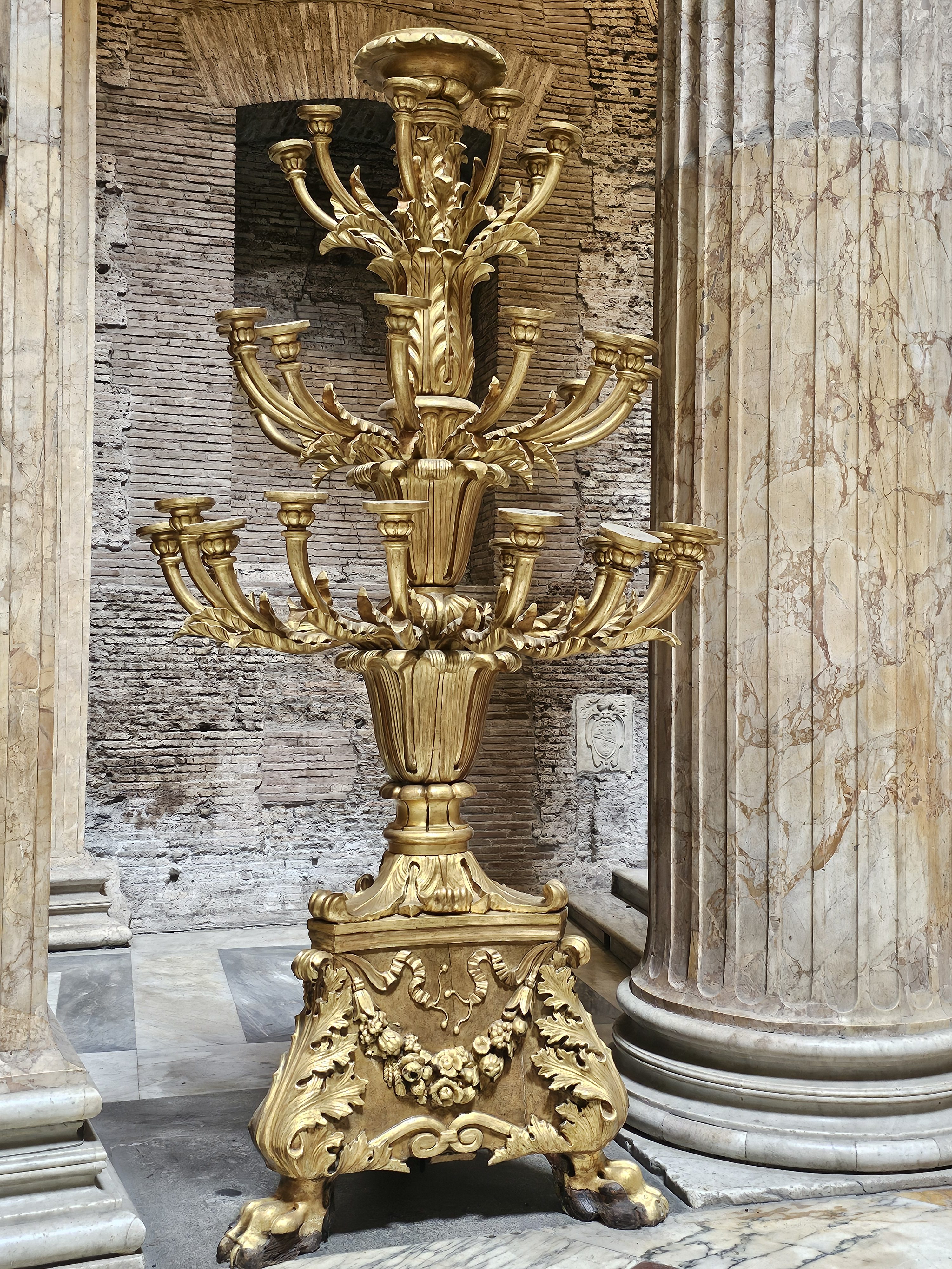 A large, ornate, golden candelabra. It is positioned indoors, likely within a historical building, given the marble columns and brick wall visible in the background. Captured from the Pantheon in Rome, Italy.
