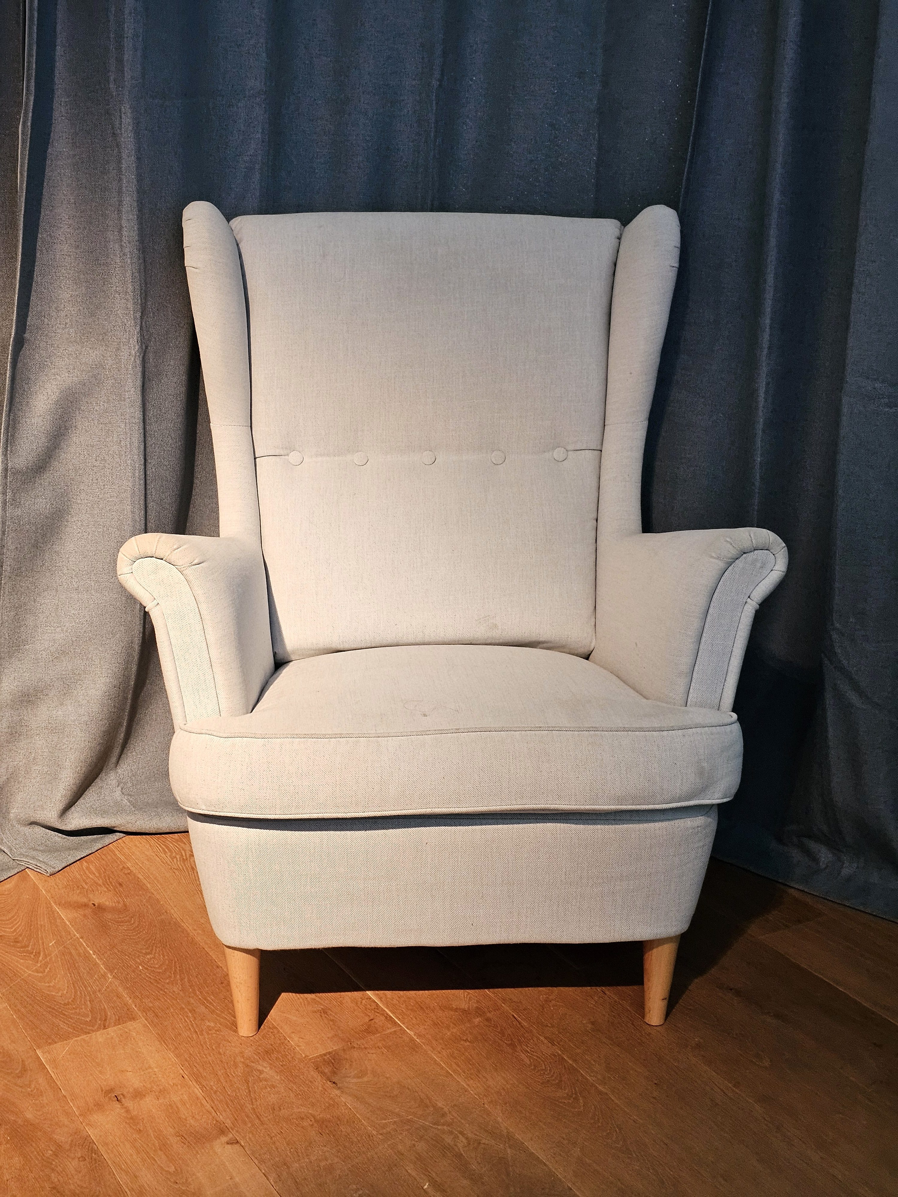 A beige wing chair, featuring a high back, armrests, and four wooden legs. It is positioned centrally against a backdrop of draped gray curtains and a wooden floor. Captured from Salzburg, Austria.