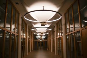 A hallway with wooden-framed glass walls on both sides, featuring large circular ceiling lights hanging in a row. The corridor appears empty and leads to a set of double doors at the end.