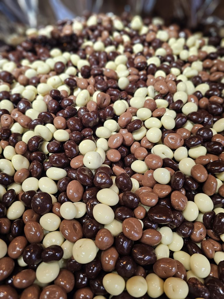 A close-up view of a pile of chocolate-covered coffee beans. The beans are coated in three different types of chocolate: dark chocolate, milk chocolate, and white chocolate.