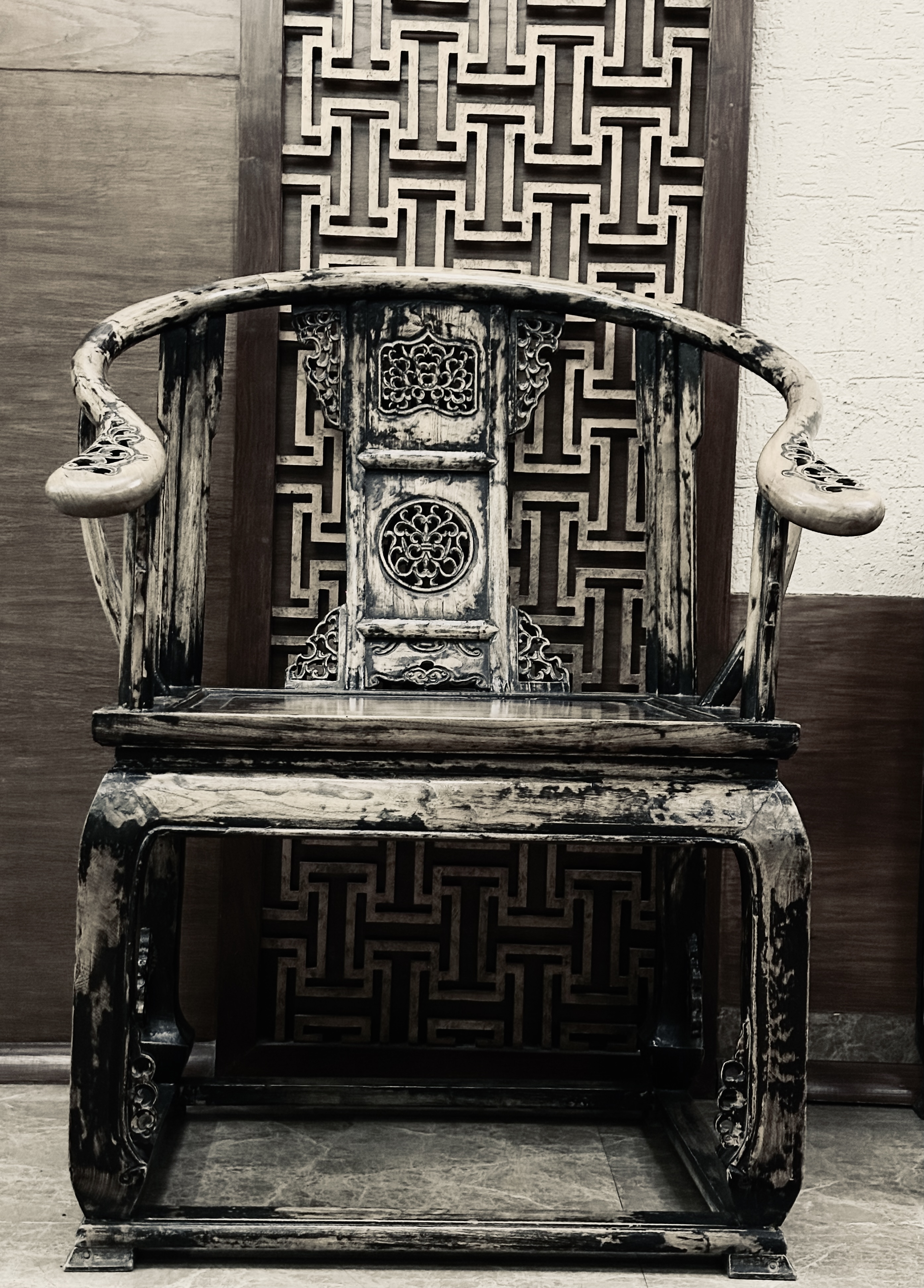 Vintage wooden chair with intricate carvings on the backrest and arms, set against a decorative wall with a geometric pattern.