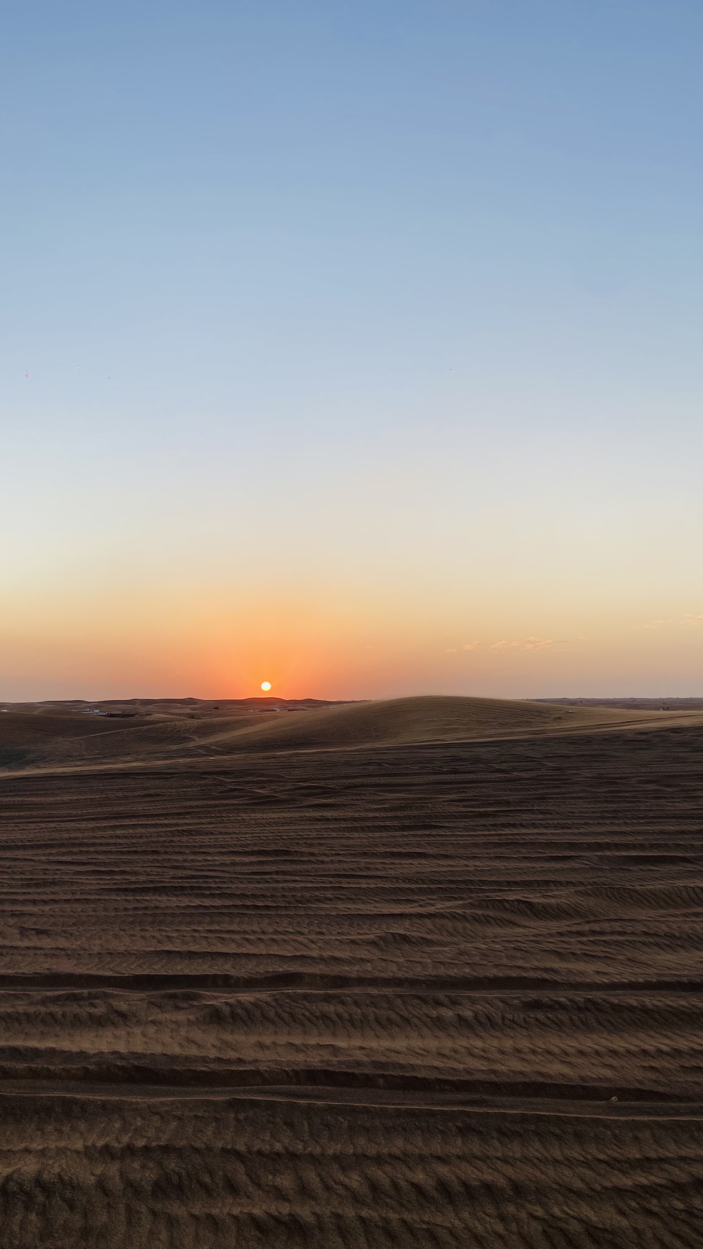 A serene desert landscape at sunset with the sun setting on the horizon, casting a warm glow over the sand dunes and a clear blue sky above.