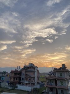 A picturesque view of a suburban area at sunset, featuring two multi-story houses with distinct architectural designs. The sky is filled with soft, scattered clouds, illuminated by the warm glow of the setting sun, while mountains are visible in the background.