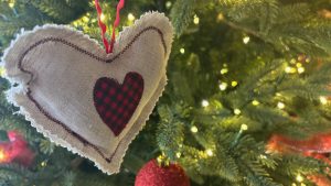 Closeup of an ornament made of cloth, shaped like a heart, in the branches of an evergreen tree with lights.