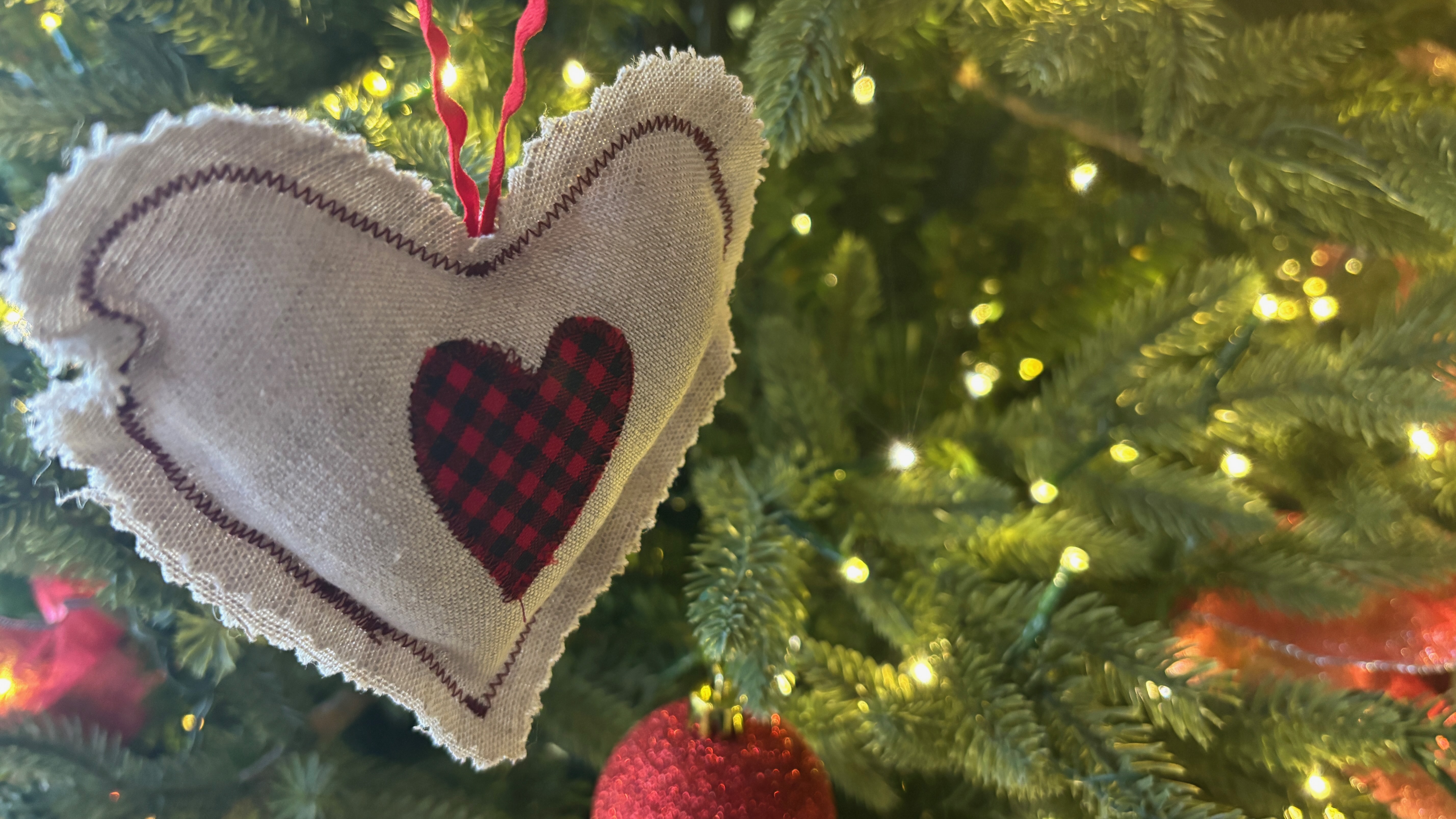 Closeup of an ornament made of cloth, shaped like a heart, in the branches of an evergreen tree with lights.