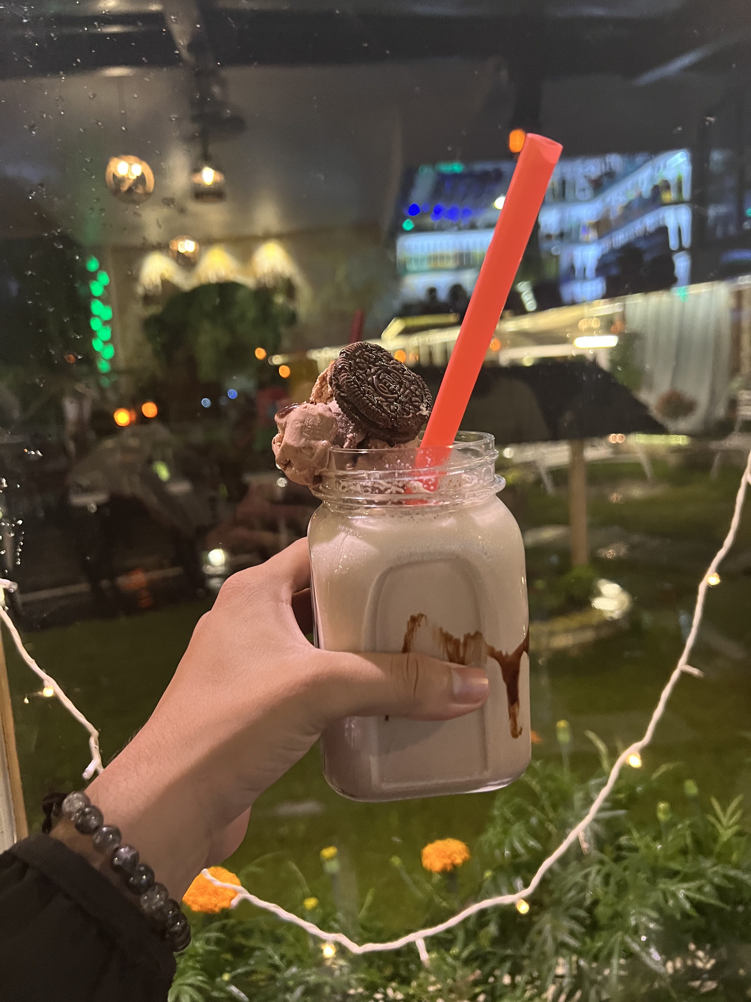 A hand holding a mason jar filled with a milkshake topped with whipped cream, a cookie, and a red straw. The background shows a dimly lit outdoor setting with decorative lights and blurred greenery.