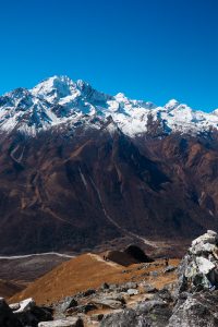 A breathtaking view of a snow-capped mountain range under a clear blue sky. In the foreground, a rocky, brown hillside with two small figures walking along a path, suggesting a hiking expedition.