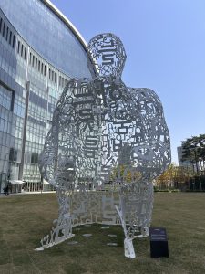 A large, white sculpture resembling a human figure, made up of various letters and symbols, stands on a grassy area in front of a modern glass building